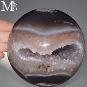 Puede incluir: Una piedra de ágata esférica pulida que presenta una superficie exterior de color marrón oscuro y translúcido con una cavidad de geoda horizontal central llena de estructuras cristalinas brillantes y capas de bandas blancas y bronceadas.