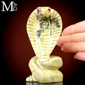Puede incluir: Una figura de cobra tallada en piedra verde claro con patrones moteados gris oscuro y pequeños ojos de color marrón rojizo. La serpiente está representada en posición erguida con una base en espiral con patrón de escamas. Las letras MAG aparecen en blanco.