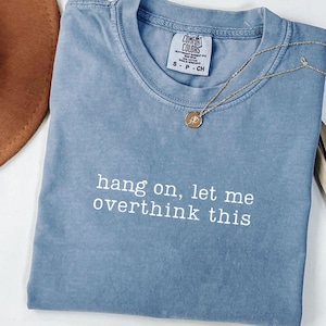 Pode incluir: Uma camiseta azul Comfort Colors com a frase branca "hang on, let me overthink this". Um colar de ouro com um pingente repousa sobre a camiseta. Um chapéu marrom é visível no fundo.