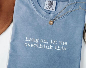 May include: A blue Comfort Colors t-shirt with the phrase "hang on, let me overthink this" printed in white. A gold necklace with a pendant rests on the shirt. A brown hat is visible in the background.