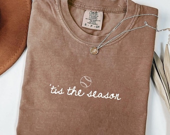May include: A brown t-shirt with the text "'tis the season" and a baseball graphic. The shirt has a crew neck and a small tag at the neckline. A silver necklace with a baseball pendant is draped across the shirt.