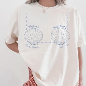 May include: Cream-colored t-shirt with a blue seashell design. The text on the shirt reads "Meghan's Bachelorette Miami, Florida est. 2024". The shirt is paired with red and white checkered shorts.