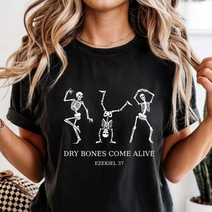 May include: A black short-sleeve t-shirt featuring a white graphic of three skeletons in various dancing poses. Below the skeletons, the text reads "DRY BONES COME ALIVE" followed by "EZEKIEL 37" in a clean, white, sans-serif font.