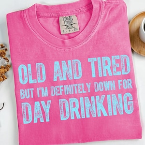 May include: A pink Comfort Colors t-shirt with the text "OLD AND TIRED BUT I'M DEFINITELY DOWN FOR DAY DRINKING" in light blue. The shirt is folded and has a small tag on the collar.