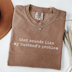 May include: A brown Comfort Colors t-shirt with the text "that sounds like my husband's problem" in white. A silver necklace with a small pendant is draped over the shirt. A brown hat is visible in the background.