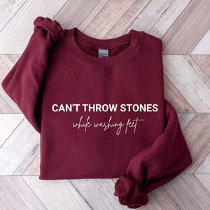 May include: A maroon sweatshirt with the text "CAN'T THROW STONES while washing feet" in white script. The sweatshirt is folded on a light brown and white wooden surface.