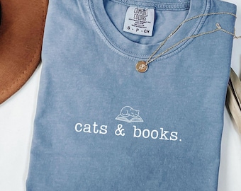 May include: A blue Comfort Colors t-shirt with the words "cats & books" in white script. A gold necklace with a circular pendant rests on the shirt. The shirt has a small graphic of a cat on a book.