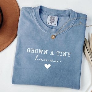 May include: A blue Comfort Colors t-shirt with the words "GROWN A TINY human" and a white heart graphic. A gold necklace with a pendant is draped across the shirt. The shirt is folded neatly on a white surface.