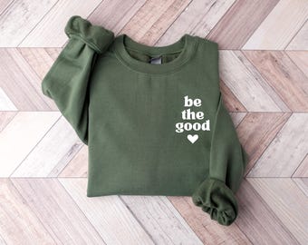 Puede incluir: Una sudadera verde oliva con las palabras "be the good" en escritura blanca y un corazón blanco. La sudadera está doblada sobre una superficie de madera marrón claro y blanca. Las mangas están dobladas.