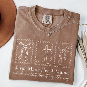 May include: A brown Comfort Colors t-shirt with the text "Jesus Made Her A Mama and she wouldn't have it any other way." The shirt features three white framed images: a bow, a cross, and another bow. A silver necklace with a pendant is draped across the shirt.