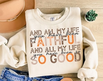 May include: Cream-colored sweatshirt with the text "AND ALL MY LIFE you have been FAITHFUL AND ALL MY LIFE you have been SO GOOD" in brown, orange, and black lettering. A tan hat and blue jeans are visible.