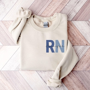 Pode incluir: Camisola creme com as letras "RN" em azul e o nome "Jennifer" em fonte cursiva. A camisola tem gola redonda e mangas compridas. A peça de vestuário é feita de um material macio.