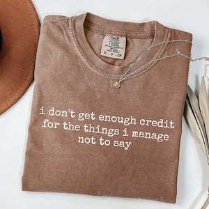 Pode incluir: Uma camiseta Comfort Colors marrom com o texto branco "i don't get enough credit for the things i manage not to say". Um colar de prata com um pequeno pingente está sobre a camiseta.
