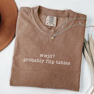 May include: A brown Comfort Colors t-shirt with the text "wwjd? probably flip tables" in white. A silver necklace with a small pendant is draped over the shirt. The shirt is folded and placed on a white surface.