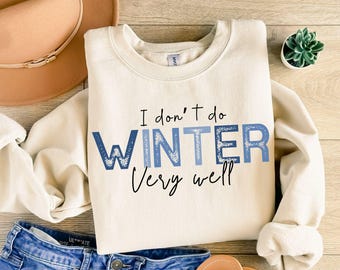 Puede incluir: Sudadera color crema con el texto "I don't do WINTER Very well" en azul y negro. La palabra WINTER está en una fuente azul con un diseño de copo de nieve. Se ven un sombrero y unos vaqueros.