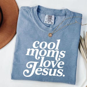 Puede incluir: Una camiseta azul Comfort Colors con el texto "cool moms love Jesus" en blanco. Un collar dorado con un colgante está drapeado sobre la camiseta. La camiseta es talla pequeña.