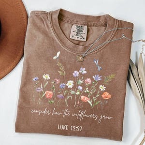 Puede incluir: Camiseta marrón con un diseño de flores silvestres y el texto "consider how the wildflowers grow" y "LUKE 12:27". Un collar plateado con un colgante está colocado sobre la camiseta. La camiseta está doblada sobre una superficie blanca.