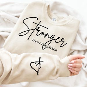 May include: A beige sweatshirt with the words "Stronger Than The Storm" in black script. The sleeve features a heart and cross design. The sweatshirt is made of a soft material.