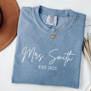 May include: A blue t-shirt with the words "Mrs. Smith" and "EST 2025" printed in white script. A gold necklace with a pendant rests on the shirt. The shirt is folded and has a tag that says "Comfort Colors".