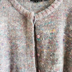 May include: A light gray knit cardigan featuring a multicolored speckled pattern with flecks of blue, pink, and yellow. The garment has a round neckline, a button-down front with matching light gray buttons, and a label reading Jeanne Pierre.