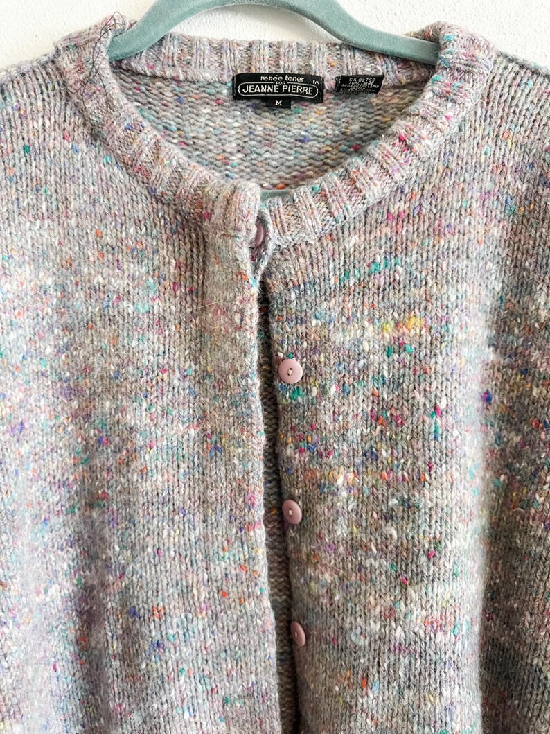 May include: A light gray knit cardigan featuring a multicolored speckled pattern with flecks of blue, pink, and yellow. The garment has a round neckline, a button-down front with matching light gray buttons, and a label reading Jeanne Pierre.