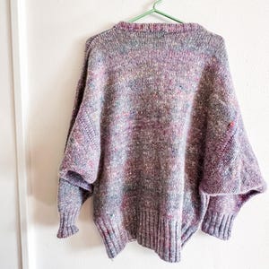 May include: A long-sleeved knit sweater featuring a multi-colored speckled pattern with dominant shades of purple, blue, and pink. The garment has a ribbed crew neckline, ribbed cuffs, and a ribbed hem, with a relaxed fit and dropped shoulder seams.