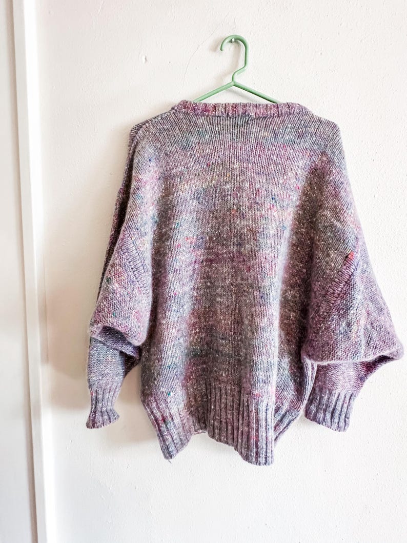 May include: A long-sleeved knit sweater featuring a multi-colored speckled pattern with dominant shades of purple, blue, and pink. The garment has a ribbed crew neckline, ribbed cuffs, and a ribbed hem, with a relaxed fit and dropped shoulder seams.