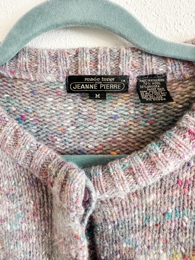 May include: A light gray knit cardigan with a multicolored speckled pattern, featuring a ribbed crew neckline and a button-front closure. The interior label reads Renee Tener for Jeanne Pierre, size M, made of 70% wool and 30% angora rabbit hair.