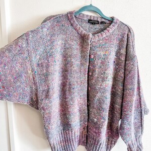 May include: A long-sleeve knit cardigan in a heathered lavender tone with multicolored flecks of pink, blue, and yellow throughout. The garment features a round neckline, a button-down front with light pink buttons, and ribbed cuffs and hem.