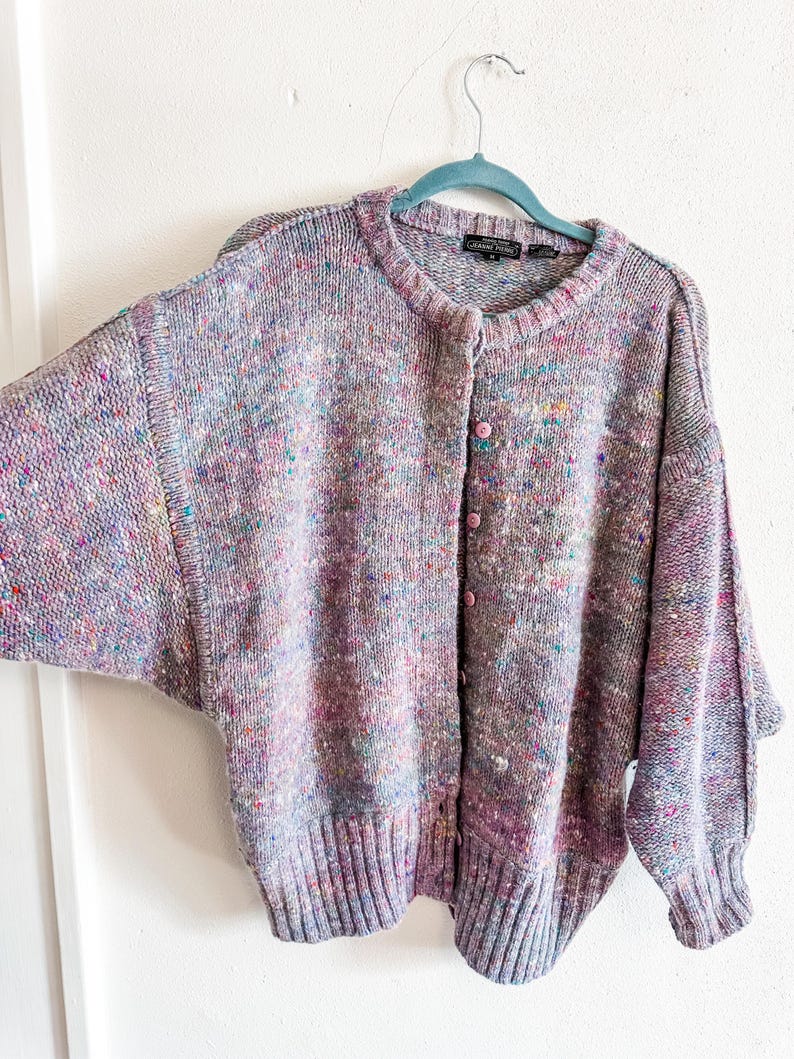 May include: A long-sleeve knit cardigan in a heathered lavender tone with multicolored flecks of pink, blue, and yellow throughout. The garment features a round neckline, a button-down front with light pink buttons, and ribbed cuffs and hem.