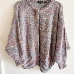 May include: A long-sleeve button-up cardigan in a heathered lavender knit with multicolored flecks of blue, yellow, and pink. The garment features a ribbed crew neckline, ribbed cuffs, a ribbed hem, and a black label reading Jeanne Pierre.