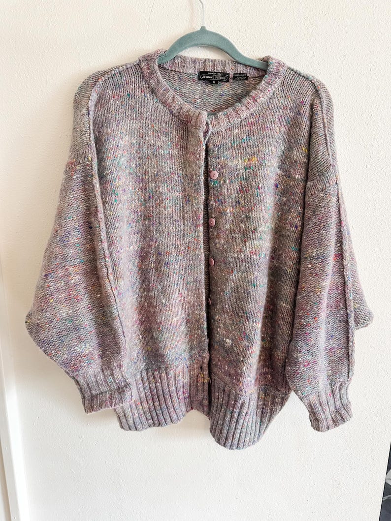 May include: A long-sleeve button-up cardigan in a heathered lavender knit with multicolored flecks of blue, yellow, and pink. The garment features a ribbed crew neckline, ribbed cuffs, a ribbed hem, and a black label reading Jeanne Pierre.