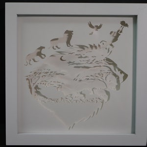 May include: White paper cut-out art framed in a white frame. The art depicts a landscape with a moon, trees, and animals including a lion, a bear, and birds.