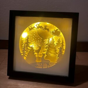 Könnte beinhalten: Eine gerahmte, beleuchtete Weihnachtsszene. Das Kunstwerk zeigt ein kreisförmiges Design mit Silhouetten von Hirschen, Bäumen und dem Schlitten des Weihnachtsmanns vor einem gelben Hintergrund. Der Rahmen ist schwarz, und der Gesamteffekt ist ein warmes, festliches Leuchten.