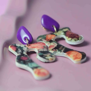 Op de afbeelding: Een paar oorbellen met bloemenprint en paarse accenten. De oorbellen zijn gevormd als abstracte vormen met een witte achtergrond en een bloemenpatroon.