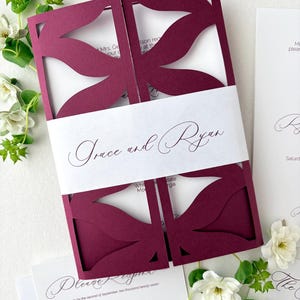May include: A deep burgundy laser-cut gatefold wedding invitation featuring a floral-inspired cutout pattern. A white paper belly band wraps around the centre, printed with the names Grace and Ryan in a black script font, accompanied by matching white stationery inserts.