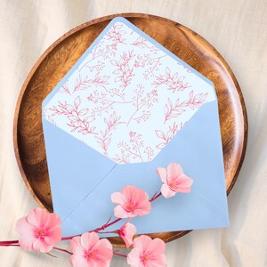 May include: A pale blue envelope with a floral design inside, presented on a wooden plate. Pink flowers and a branch are in the foreground. Suitable for stationery and invitations, adding a touch of elegance.