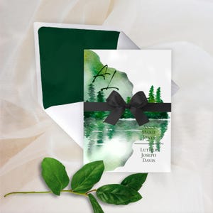 May include: A wedding invitation with a watercolour landscape design featuring a lake and trees, accented with a black ribbon. The invitation includes the names Anna Marie Jones and Luther Joseph Davis. The envelope is dark green.