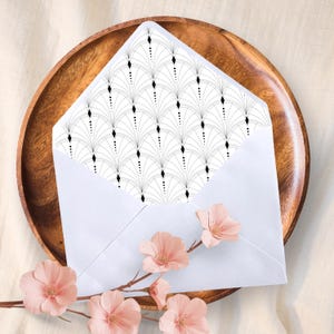 Könnte beinhalten: Ein weißer Umschlag mit einem schwarz-weißen Art-Deco-Muster im Inneren, der auf einem Holztablett liegt. Rosa Blüten mit braunem Stiel befinden sich im Vordergrund und verleihen der Szene einen Farbtupfer.