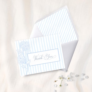 May include: A white thank you card with a blue floral design and the words "Thank You" in cursive. The card has a blue striped background and is accompanied by a matching envelope. The card and envelope are on a white surface.