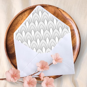 May include: A white envelope with a black and white geometric design inside, presented on a wooden plate. Pink flowers and a branch are placed near the envelope, creating a decorative arrangement.