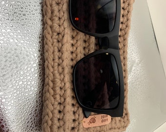 May include: Black sunglasses rest on a tan crocheted case. The case is rectangular and has a textured appearance. A small, rectangular tag with the words "HAND MADE" is attached to the case.