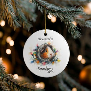 Könnte beinhalten: Ein rundes, weißes Keramikornament mit einer Aquarellillustration eines Meerschweinchens, das in bunten Lichterketten verheddert ist. Der Text "SEASON'S" steht über dem Meerschweinchen und das Wort "Squeakings" ist darunter in Schwarz gedruckt.