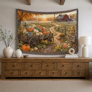 May include: A woven wall tapestry featuring a rural autumn landscape with a wooden wagon overflowing with pumpkins. The scene includes a red barn, a dirt path, and a wooden sign with an arrow pointing toward the text PUMPKINS. The tapestry has a fringed edge.