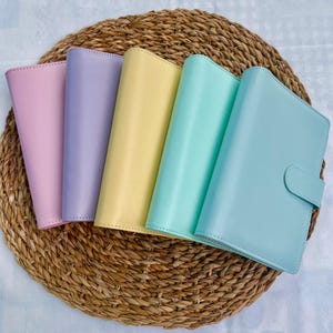 May include: Five leather-bound notebooks in different colours arranged in a semi-circle on a woven straw mat. The notebooks are pink, lavender, yellow, light blue, and a darker shade of blue.