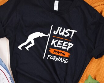 May include: Black t-shirt with a white silhouette of a person pushing against a wall. The text "JUST KEEP MOVING FORWARD" is printed in orange on the shirt.
