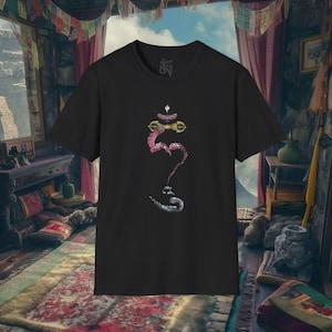 May include: Black t-shirt featuring a colorful, stylized illustration of a spiritual symbol. The design incorporates pink, gold, and silver elements. The shirt is displayed in a room with a rug and curtains.