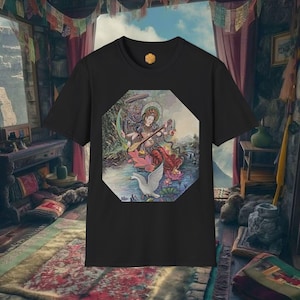 Puede incluir: Camiseta negra con una colorida ilustración de una deidad tocando un instrumento de cuerda, sentada sobre una flor de loto, con un cisne. La obra de arte está enmarcada en forma octogonal. La camiseta tiene cuello redondo y mangas cortas.