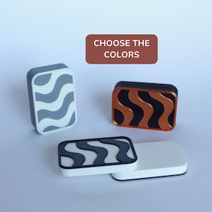 Op de afbeelding: Drie rechthoekige tegels met een golvend patroon. De tegels zijn in grijs en wit, zwart en koper, en zwart en wit. De tekst "CHOOSE THE COLORS" staat op een rode rechthoek.