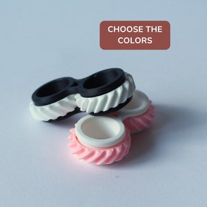 May include: Three fidget spinners in black, white, and pink. The spinners are stacked on top of each other. The top spinner is black, the middle spinner is white, and the bottom spinner is pink.